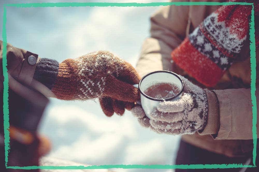 One person giving a warm tea to the other person, both clothed in warm wool clothes and mittens, on a cold winter day, indicating an article on 5 rare herbs that you must have in your winter apothecary to protect your health during wintertime.